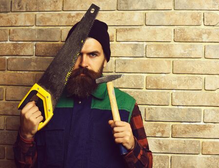 bearded worker man holding saw and hammer with serious faceの写真素材