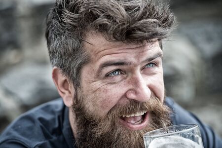 Man with beard and mustache drinks beer, defocused background. Masculinity concept. Hipster with tousled hair and gray, guy with wrinkles on cheerful face drinking beverage out of glass.の写真素材