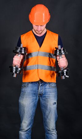 Man in helmet, hard hat holds heavy dumbbells, black background. Worker, contractor, builder on busy...