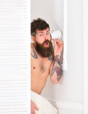 Hipster  on stunned face secretly listen conversation. Lovers and cheating concept. Man with beard and mustache eavesdrops using mug near wall. Man in white interior spying, eavesdroppingの写真素材