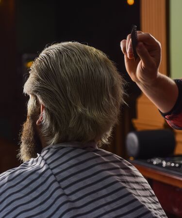 Guy with long dyed blond hair close up. Do not cut your own hair. Barber works on hairstyle for bearded man barbershop background. Hipster client getting haircut. Barber with scissors and client.の写真素材