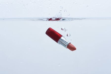 Water resisting red lipstick fall into pure, transparent water, white background. Water resistance cosmetic concept. Lipstick under water with trail of transparent bubbles, copy space.の写真素材