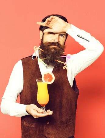 handsome bearded man with colorful tubes in long beard and mustache has stylish hair on serious face holding glass of alcoholic cocktail in vintage suede leather waistcoat, on red studio backgroundの写真素材