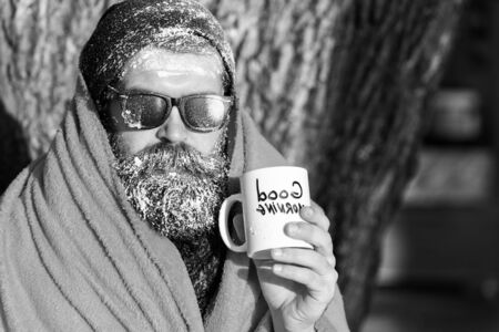 Frozen man, bearded hipster, with beard and moustache in black sunglasses covered with white frost wrapped in orange blanket with warming drink in cup on winter day outdoors on natural backgroundの写真素材