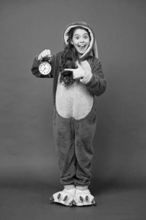 Counting time. It is weekend. Adorable bunny hold alarm clock. Small girl in bunny costume. Child in rabbit kigurumi. Happy girl bunny pajamas. Rest and relax. Bunny kid red background. Baby animalの写真素材