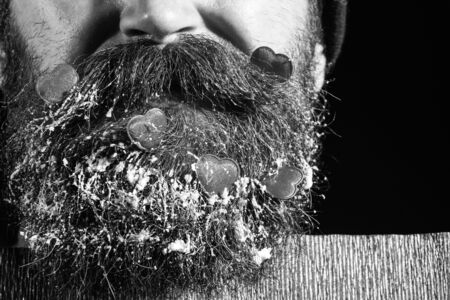 handsome bearded man or guy with fashionable mustache, has closed mouth on face and red decorated hearts with snow in beard near paper on black background, copy spaceの写真素材