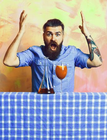 Bearded man, long beard. Brutal caucasian tattooed surprised hipster with moustache in denim shirt with tropical alcohol fresh cocktail and flask on blue checkered plaid on colorful texture backgroundの写真素材
