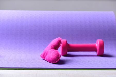 Barbell and pink love symbol, close up. Love of sports and fitness idea. Healthy shape concept. Dumbbell made of pink plastic near soft toy heart on purple texture background, copy spaceの写真素材