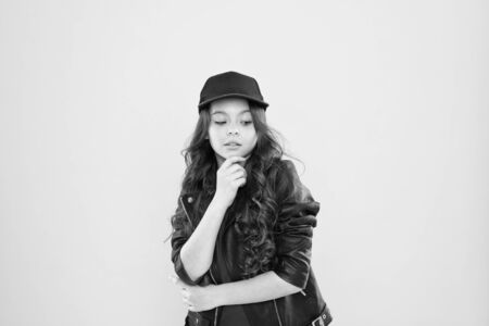 how to be a hipster. happy kid in stylish leather jacket. small girl just thinking. kid fashion concept. child has long curly hair. after visit to hairdresser. better look in cap. looking so coolの写真素材