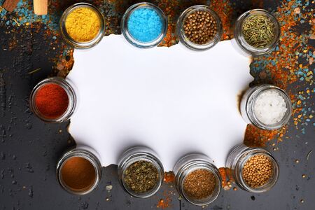 Glass jars with paprika, mustard, oregano and other spices near granulated sea salt. Food art concept. Composition of bottles with herbs around paper piece. Set of spices on dark grey wood backgroundの写真素材