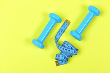 Shaping and fitness equipment. Sports and healthy lifestyle concept. Dumbbells and measure tape in cyan blue color on light yellow background, top view. Barbells next to roll of tape measureの写真素材