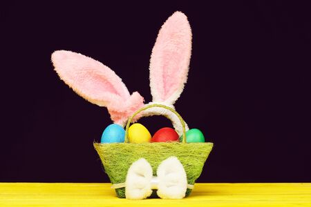 Rabbit ears in pink colour put into green basket full of painted Easter eggs and white fluffy bow...