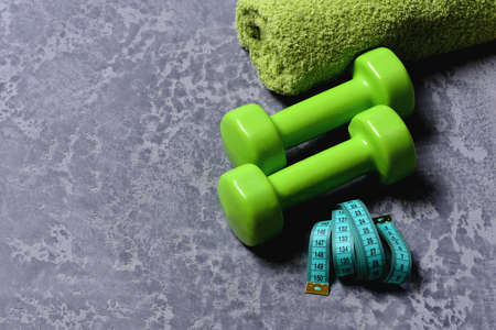 Workout and measurement concept. Sports and healthy training idea. Measuring tape next to plastic barbells. Dumbbells in green color, towel and cyan tape roll on grey texture background, top viewの写真素材
