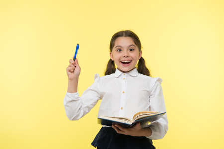 Girl cute schoolgirl in uniform hold book or textbook yellow background. Diligent pupil get knowledge from book. Child wear school uniform prepare for lesson her knowledge. Excited about knowledgeの写真素材