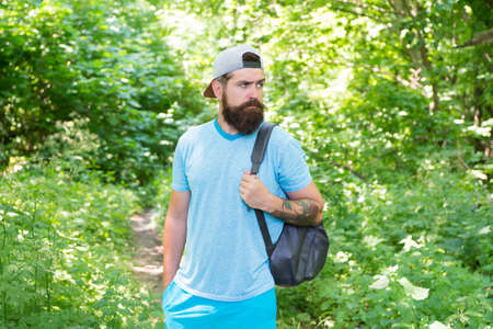 bearded man look confidently. brunette bearded man wear cap in forest on background of trees. brutal brunette bearded man with backpack. mature guy with beard in wood. nice summer dayの写真素材