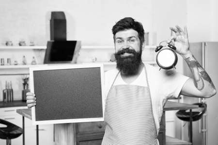 happy man with blackboard and alarm clock. time for meal. healthy food and drinks. male housekeeper in kitchen. cooking by recipe. bearded hipster offer menu. restaurant staff. copy spaceの写真素材