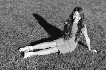 Vacation ideas. Happy smiling girl enjoy relax on lawn. Cheerful schoolgirl. Have fun. Girl cute kid sit green grass. Emotional happy kid relaxing outdoors. What makes child happy. Summer vacationの写真素材