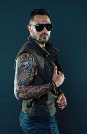 Tattooed man with biceps and triceps. Tattoo model with beard on unshaven face. Bearded man with tattoo on strong arms. Fashion macho in trendy sunglasses. Fashion style and trendの写真素材