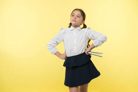 Girl cute schoolgirl in uniform hold book or textbook yellow background. Diligent pupil get knowledge from book. Child wear school uniform prepare for lesson her knowledge. Confident in her knowledgeの写真素材