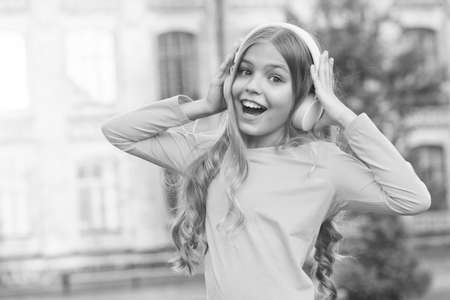 Girl listening music modern gadget. Kid happy with wireless headset outdoors sunset urban background. Stereo headphones. Kid using modern technology. Modern child. Music channel. Music for soulの写真素材