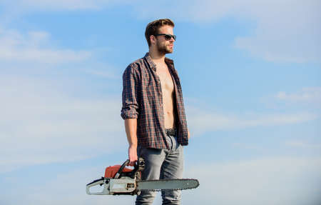 Gardener lumberjack equipment. Lumberjack with chainsaw in his hands. Dangerous job. Feeling confident. Risky temper. Powerful chainsaw. Sharp blade. Handsome man with chainsaw blue sky backgroundの写真素材