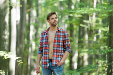enjoy weather. young unshaven man has bare torso outdoor. male beauty and fashion. guy with bristle wear checkered shirt. casual fashion style. muscular man in forest.   macho in unbuttoned shirtの写真素材