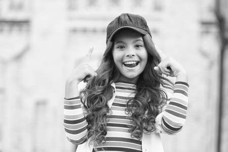 Use your brain. Happy child point fingers up outdoors. Small child wear cap on long wavy hair. Little child smile in casual wear. Fashion accessory. Trendy style. Child care and childhoodの写真素材