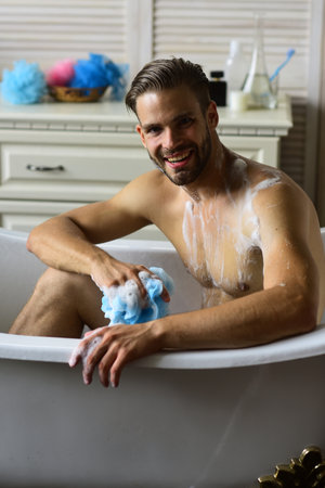 Man sitting naked in bathtub, selective focus. Relaxation concept. Guy in bathroom with toiletries on background. Man with beard and happy face taking bath.の写真素材