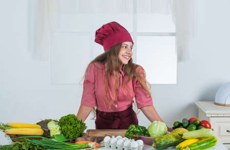 happy child cooking in kitchen. healthy and organic food only. full of vitamins. kid wear chef uniform and hat. teen girl preparing meal. fresh raw vegetables. time to eatの写真素材