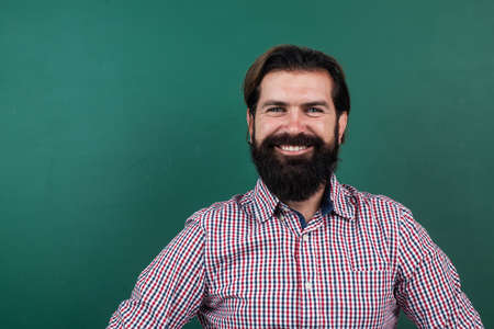 cheerful mature teacher on background of blackboard. brutal bearded man wear casual checkered shirt and smiling. express positive human emotions. caucasian hipster on green background. copy spaceの写真素材