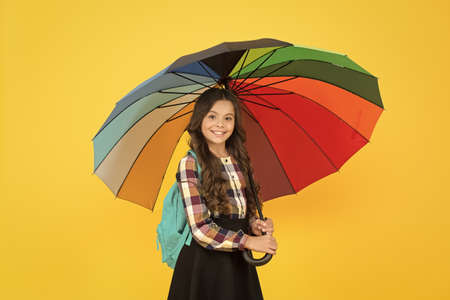 Rainy weather with proper garments. On way to school. Cheerful smiling schoolgirl. Rainy day fun. Happy walk under umbrella. Enjoy rain concept. Kid girl happy hold colorful rainbow umbrellaの写真素材