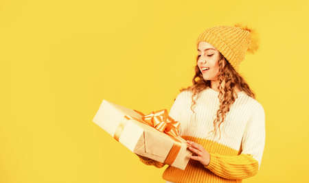 Feast of Christmas. may your christmas be bright. happy winter holidays. cheerful little girl knitted hat and sweater. kid hold present box yellow background. xmas gift shopping sale. copy spaceの写真素材