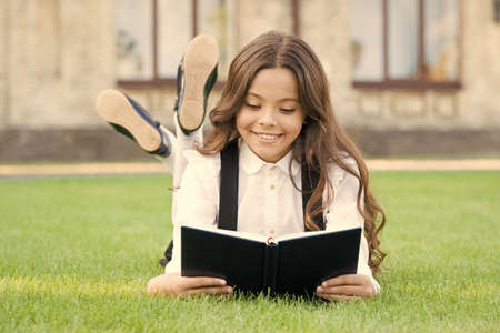 Basic education. Adorable little girl learn reading. Schoolgirl school uniform laying on lawn with favorite book. Studying concept. Extracurricular reading. Cute small child reading book outdoorsの写真素材