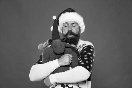 Christmas prep concept. Hipster bearded face winter sweater. Winter holiday. Soft toy. Hug and love. Buy gifts. Winter sale. Winter feast. Reindeer. Man santa hat hold christmas gift red backgroundの写真素材