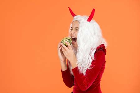 Just hanging out. happy halloween devil girl. childhood. hungry child in imp horns. kid has white hair bite pumpkin. jack o lantern. carnival costume party. trick or treat. celebrate the holidaysの写真素材