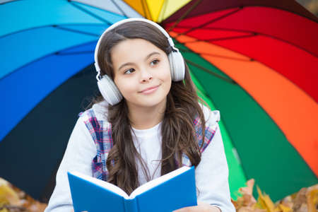 happy girl in casual style spend time listening music and reading book in autumn park enjoying good weather under colorful umbrella, back to schoolの写真素材