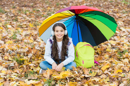 happy kid listen music in headphones while sitting in autumn forest among fallen leaves with colorful umbrella and school backpack, back to schoolの写真素材