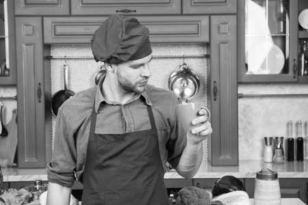 But first coffee. Man with holds coffee cup. Cook stands near table with ingredients and kitchenware. Self care. Cooking process concept. Chef in burgundy uniform drink coffee for inspirationの写真素材