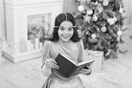 Must read for Christmas season. Happy small child read book by xmas tree. Adorable little girl read...