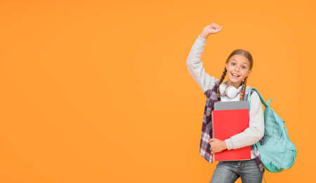 Happy girl with raised hand yellow background. Small girl ready back to school. Little girl celebrate September 1. Cute girl smile with school bag and books. Startupの写真素材