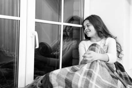 Lets get cozy. Little child feel cozy at home. Small girl sit on window sill. Cute kid rest covered in cozy plaid blanket. Cozy home. Leisure and relaxationの写真素材