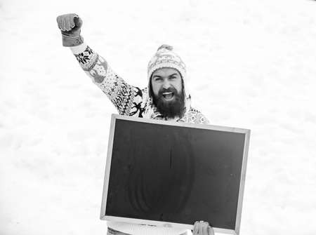 Winter event. Man with beard hold chalkboard copy space. Check it out. Winter holidays. Emotional guy snowy nature background. Winter announcement. Hipster knitted hat and gloves show blackboardの写真素材