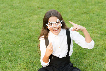 Happy child in school uniform point index finger at star-shaped glasses party accessory sitting on...