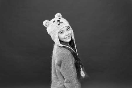 small child ready for winter. happy little girl in earflap hat. holiday activity outdoor. seasonal health care. kid fashion. Warm knitting tips. Small girl smile in knitted hat on red backgroundの写真素材