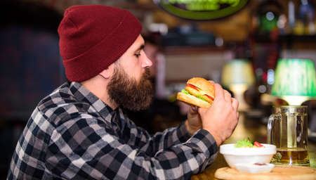 Hipster relaxing at pub. Pub is relaxing place to have drink and relax. Man with beard drink beer eat burger menu. Enjoy meal in pub. Brutal hipster bearded man sit at bar counter. High calorie snackの写真素材
