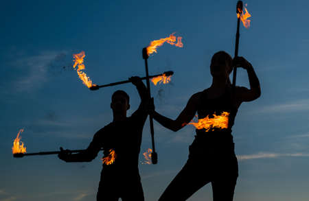 Rise to the challenge. Sexy couple manipulate burning rods. Dark silhouettes twirl fire rods on idyllic sky. Baton twirling. Fire performance. Outdoor festival. Night party. Flame and smokeの写真素材