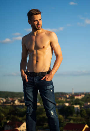 Hot day. Athletic handsome macho wear denim pants. Muscular body. Fitness model. Muscular bare torso. Six packs muscular chest. Man outdoors blue sky background. Summer trends. Male beauty conceptの写真素材