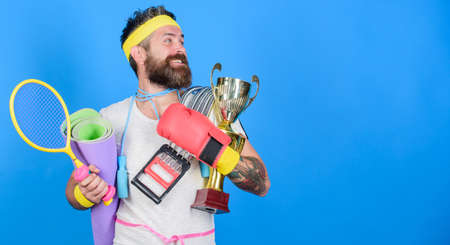 Sport concept. Sport shop assortment. Get body ready for summer. Man bearded athlete hold sport equipment jump rope fitness mat boxing glove expander racket and golden goblet. Choose sport you likeの写真素材