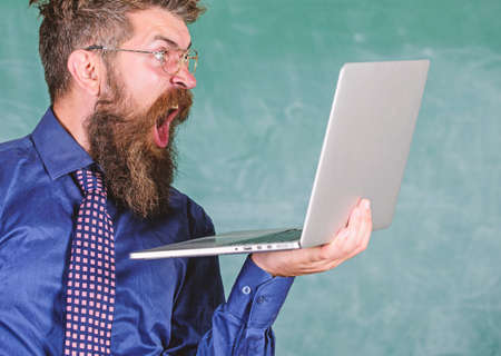 Shocking information. Hipster teacher wear eyeglasses and necktie holds laptop surfing internet. Teacher bearded shocked man with modern laptop surfing internet chalkboard background. Shocking newsの写真素材