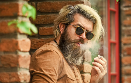 Smoking outdoors. Smoking habit. Fashionable mature man with cigarette. Brutal guy sunglasses smoking tobacco. Cool guy relaxing. Went on smoke break. Hipster smoking old architecture backgroundの写真素材